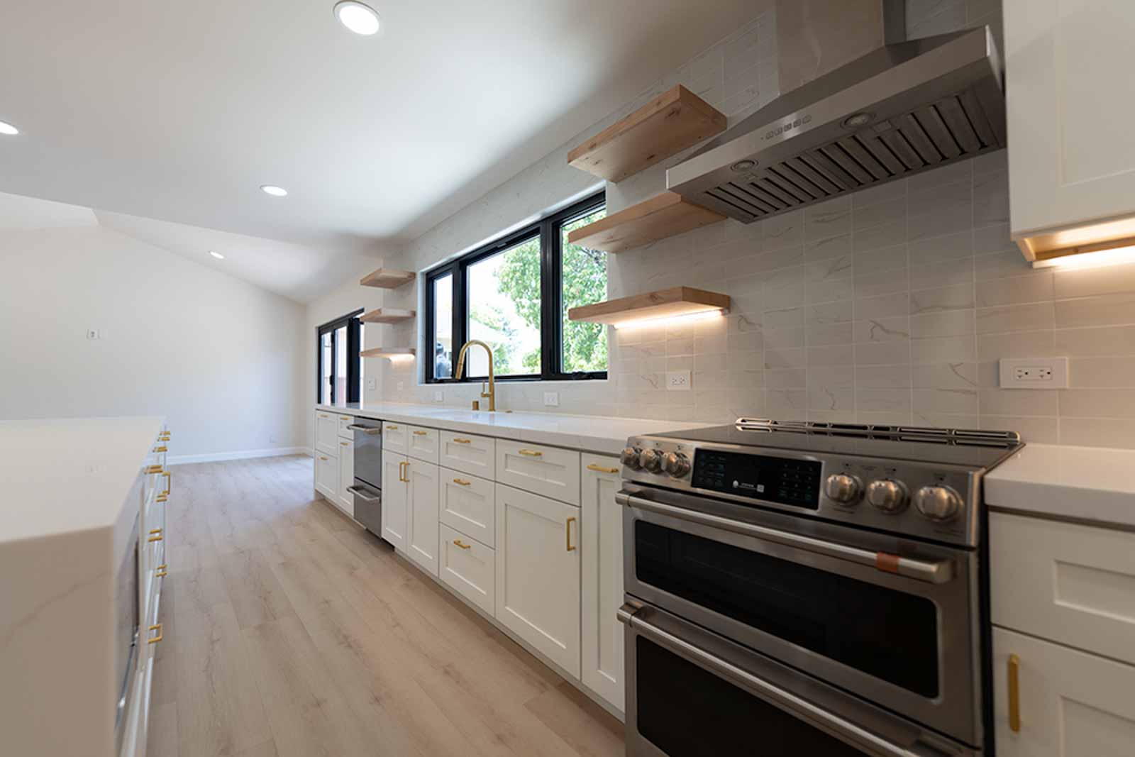 White shaker kitchen with floating wood shelves, gold faucet, stainless range, and vaulted ceiling